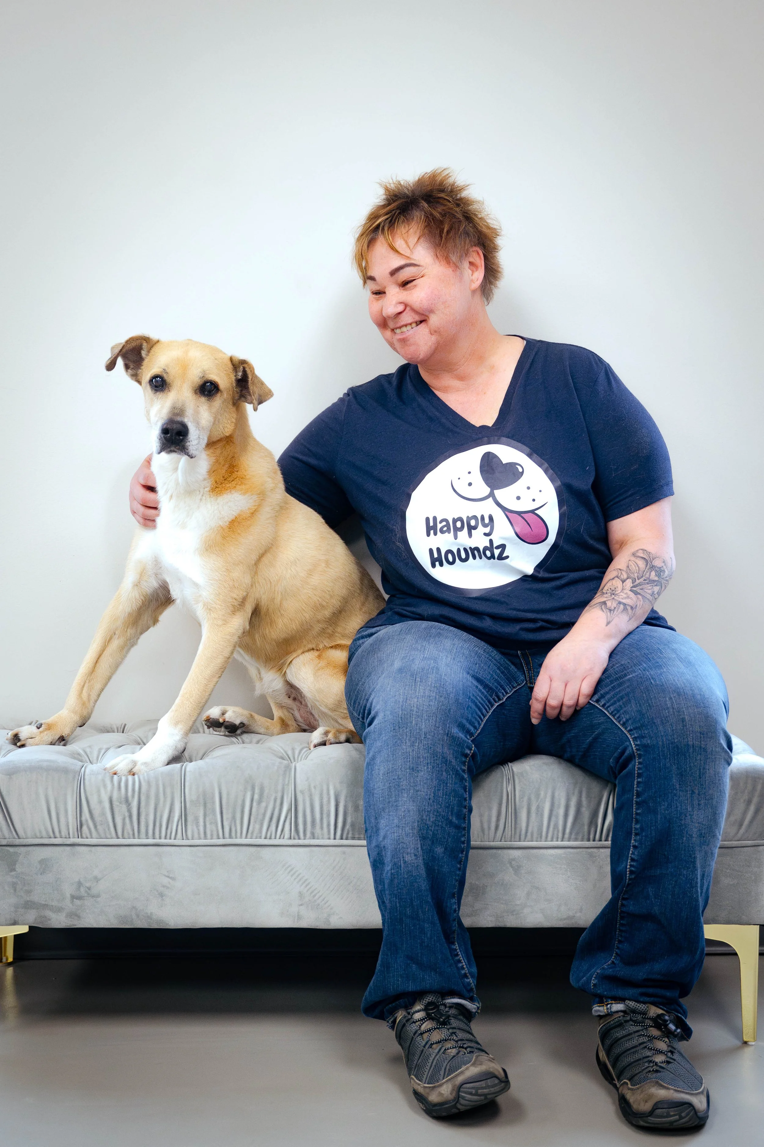 A woman sitting on a gray cushioned bench with a tan and white dog. She is wearing a navy blue t-shirt with a dog face and the words "Happy Houndz" on it, and blue jeans. The woman is smiling and has a tattoo on her left arm. The dog is sitting with one paw on the bench, looking at the camera.