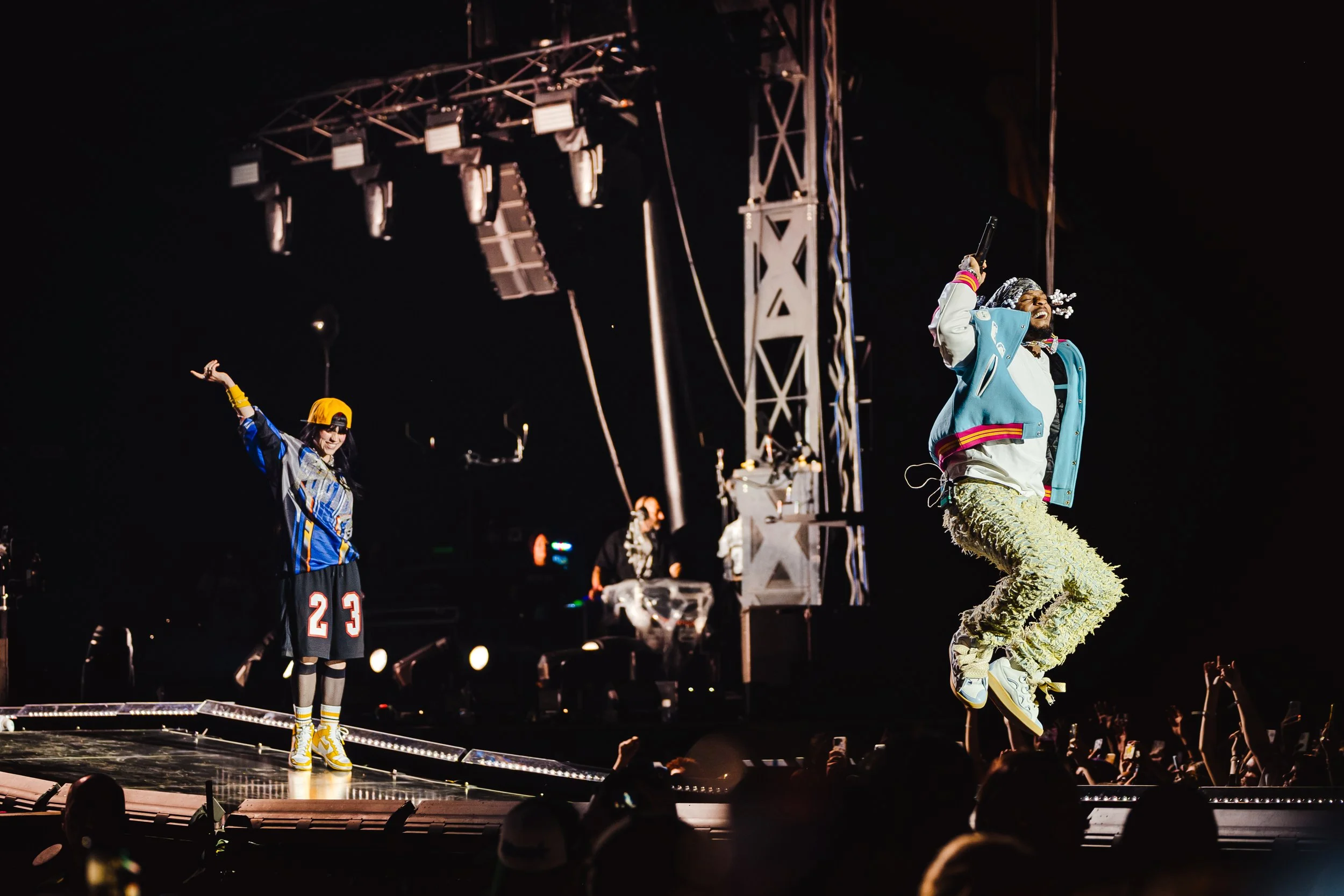 Two performers on stage at a concert, one jumping with a microphone in hand and the other standing with arms raised. Billie Eilish Armani White Osheaga Surprise special guest