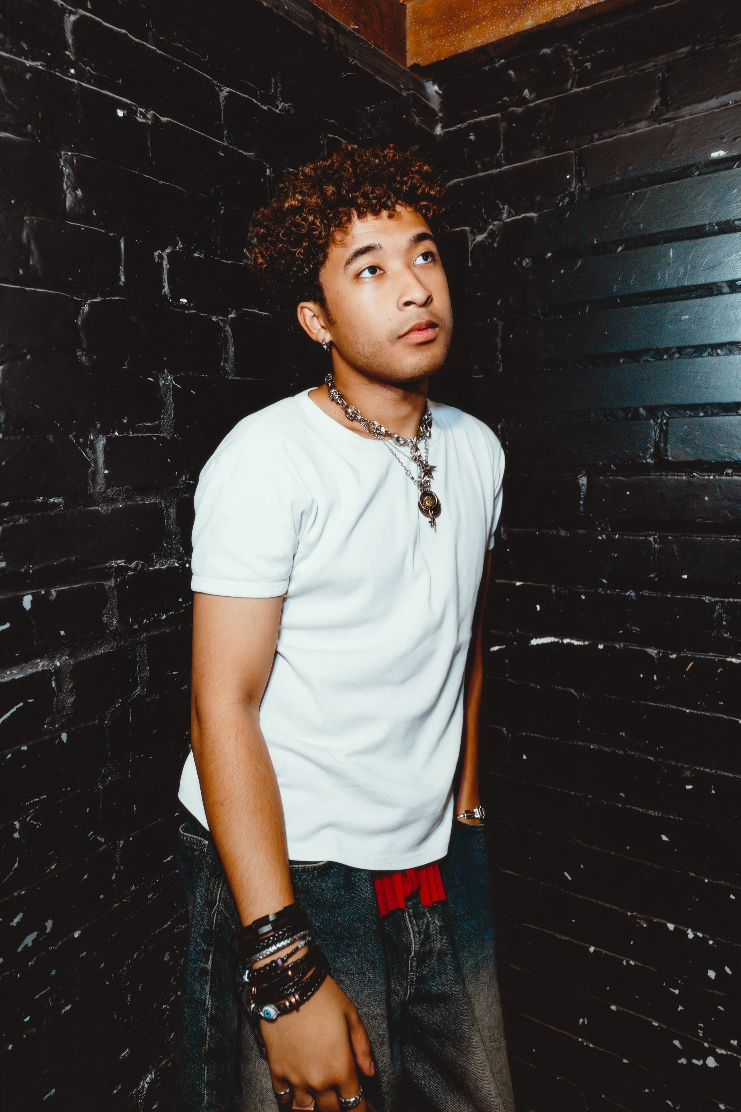 A young man with curly hair wearing a white t-shirt, multiple bracelets, and necklaces, standing against a black brick wall, looking upward.