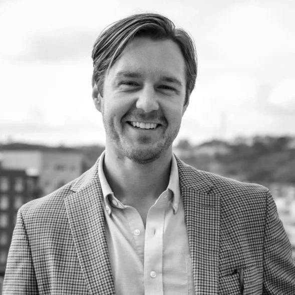 Portrait of a smiling man with light hair wearing a checkered blazer and a light-colored button-up shirt, standing outdoors with a blurred cityscape in the background.