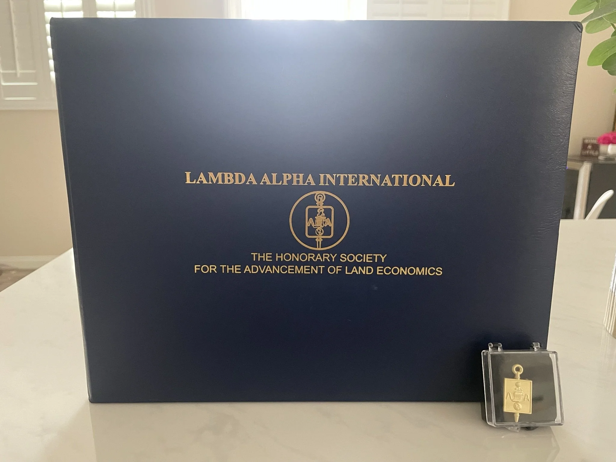 A blue box with gold text and emblem reading "Lambda Alpha International, The Honorary Society for the Advancement of Land Economics". Next to it is a small gold pin in a clear plastic case.
