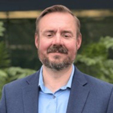 A man with a beard and short hair, wearing a blue suit and light blue shirt, standing outdoors in front of green foliage.
