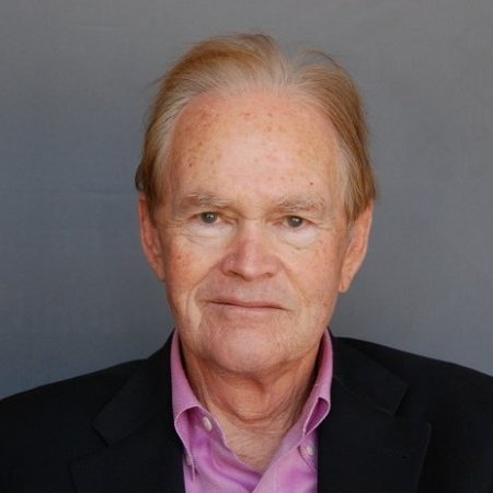 Portrait of an older man with light skin, reddish-blonde hair, wearing a black blazer and pink shirt, against a gray background.