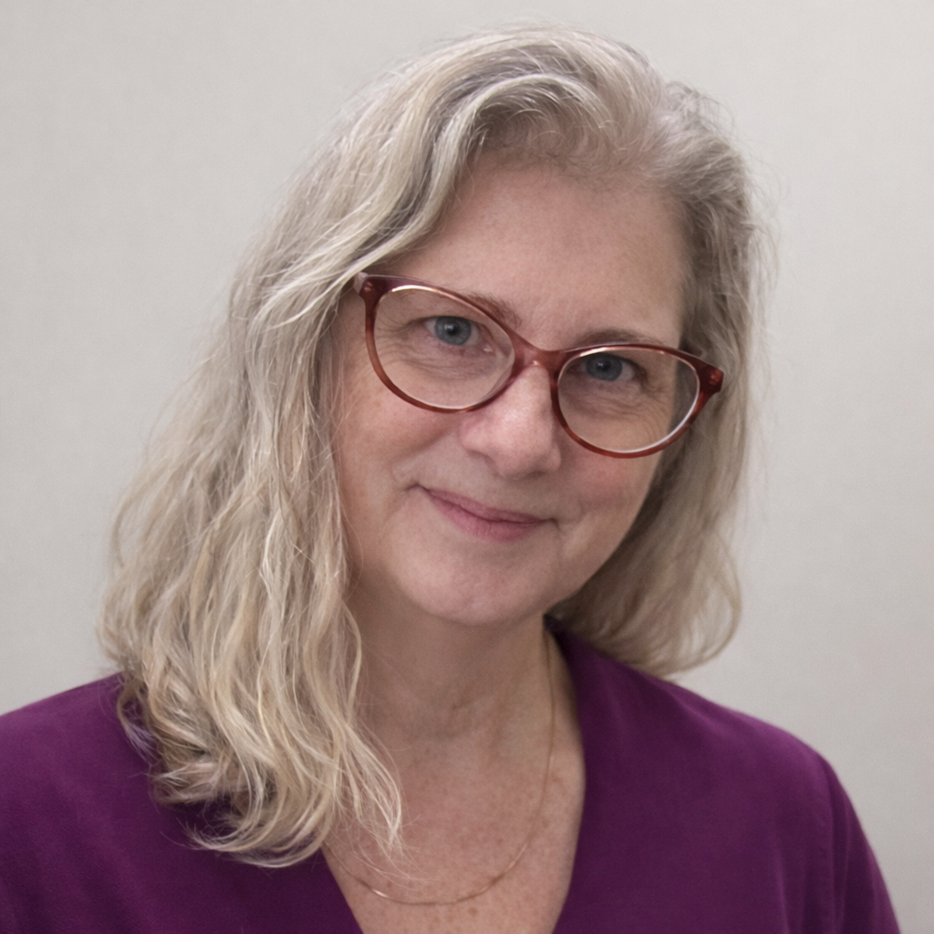A smiling woman with light blonde hair, wearing red glasses and a purple top against a plain background.
