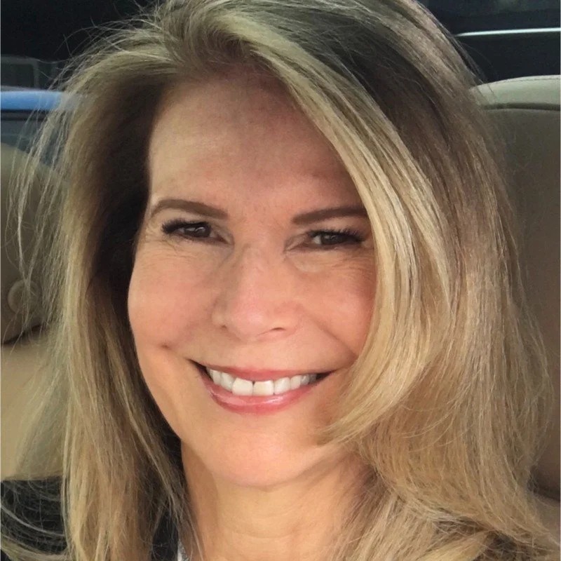A woman with long blonde hair smiling, sitting in a car.