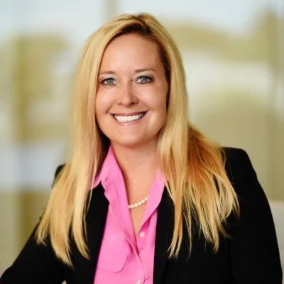 Portrait of a smiling woman with blonde hair wearing a black blazer and pink blouse indoors.