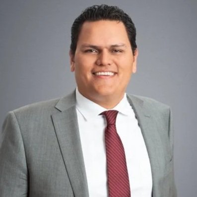 A man in a gray suit, white shirt, and red tie smiling against a gray background.