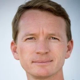 Close-up of a man with short brown hair and blue eyes against a light background.