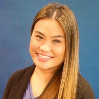 A smiling woman with long straight blonde hair, wearing a black blazer over a purple top, against a blue background.