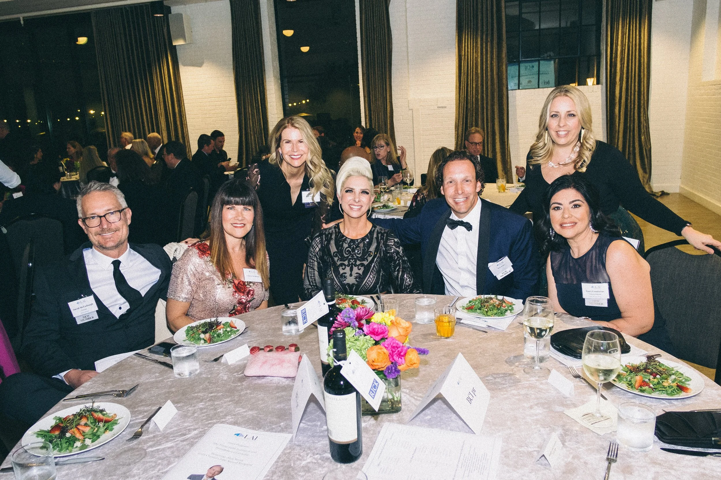 A group of eight people, five women and three men, dressed in formal attire, sitting and standing around a dining table at an event or banquet, smiling for the camera with plates of salad, glasses of wine, and a bottle of wine on the table.