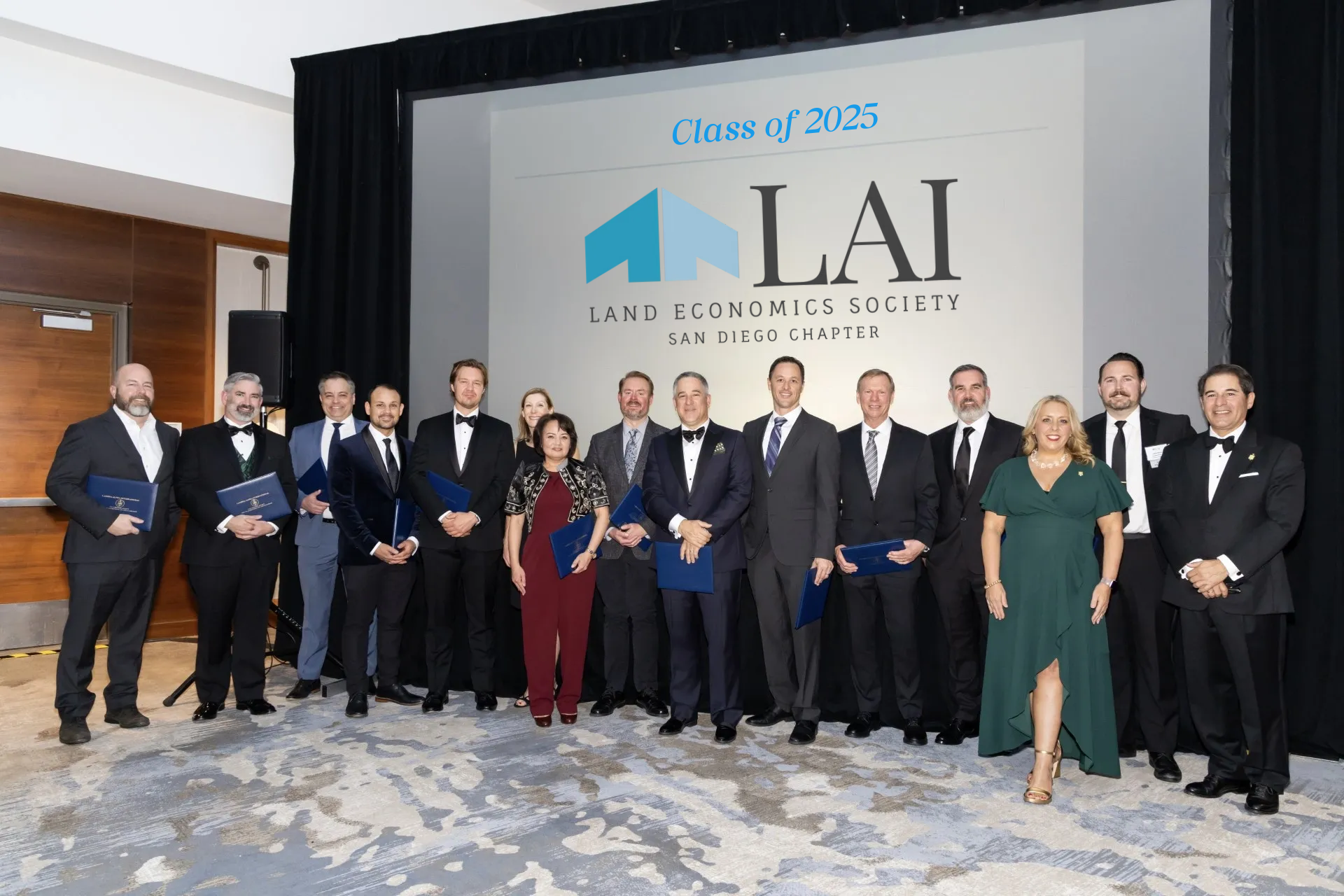 Group of people dressed in formal attire standing on stage during an event for the Land Economics Society San Diego Chapter, with a large screen behind them displaying the society's logo and the text 'Class of 2025'.