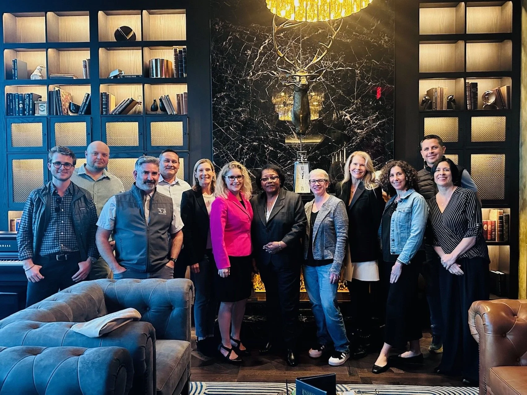 A group of fifteen people, mostly women, standing in a luxurious room with a black marble fireplace, a mounted deer head, and bookshelves. They are smiling for the photo.