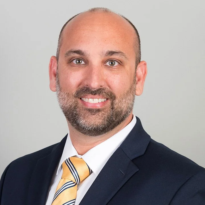 A professional headshot of a smiling man with a beard, wearing a dark suit, white shirt, and a yellow striped tie against a plain light grey background.
