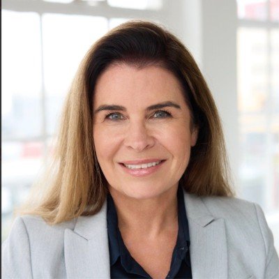 A woman with long brown hair, smiling, wearing a light gray blazer and dark blouse, standing in a bright office with large windows.