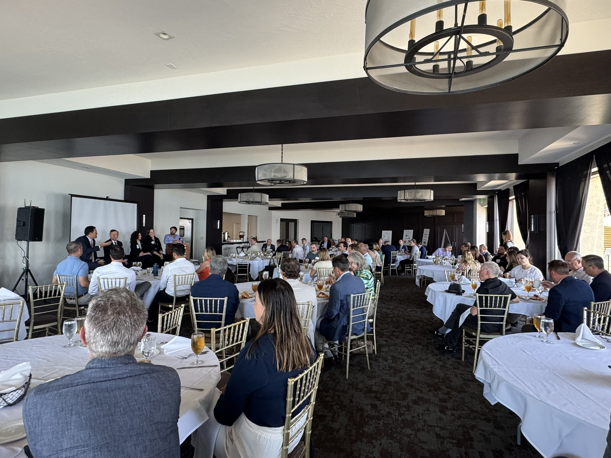 A large conference room filled with round tables of people listening to a panel discussion at the front. The room is decorated with dark wood accents and large windows with black curtains, and is illuminated by modern circular chandeliers.