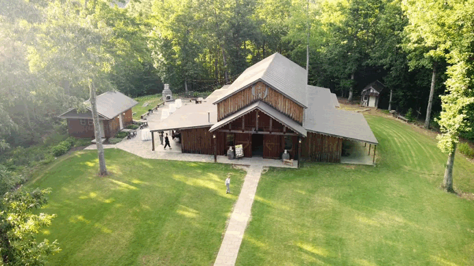A large wooden barn surrounded by green grass and trees, with a concrete walkway leading to its entrance and several people near the building.