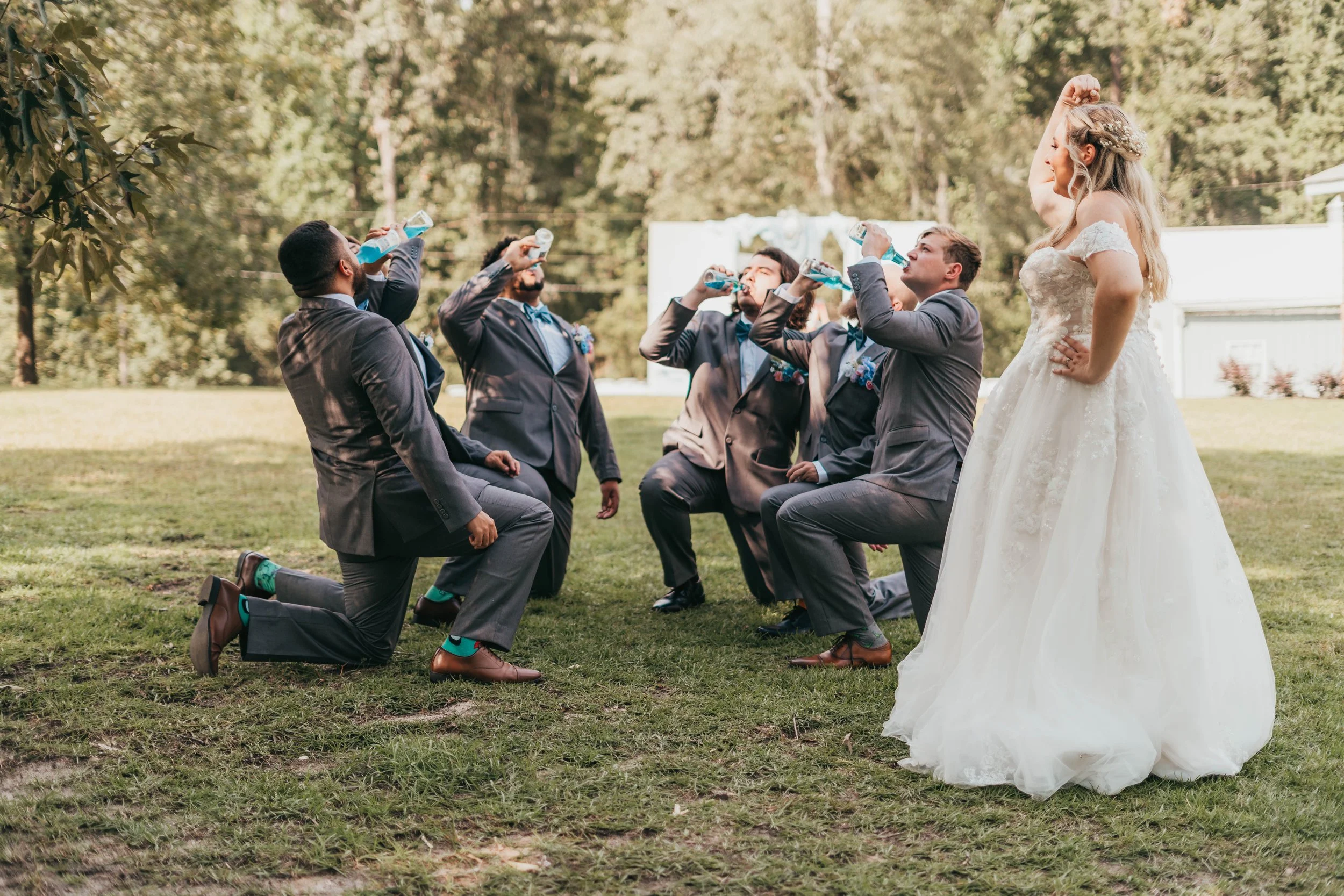Bride in a white wedding dress standing with hand on hip, surrounded by five men in suits kneeling on grass and drinking from water bottles outdoors.