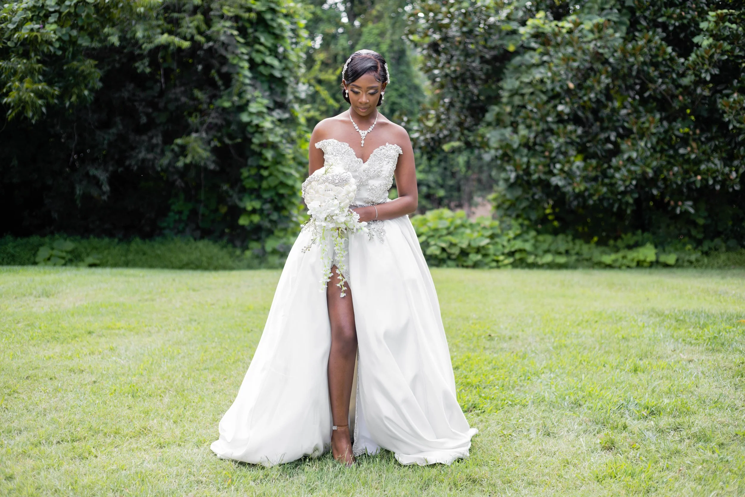 A bride in a strapless white wedding gown with a high slit, standing on grass with lush green trees in the background, holding a bouquet of white flowers.