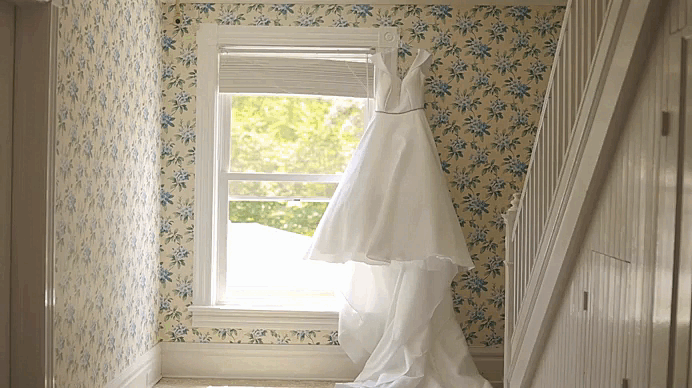 A white wedding dress hanging in front of a window in a room with floral wallpaper.