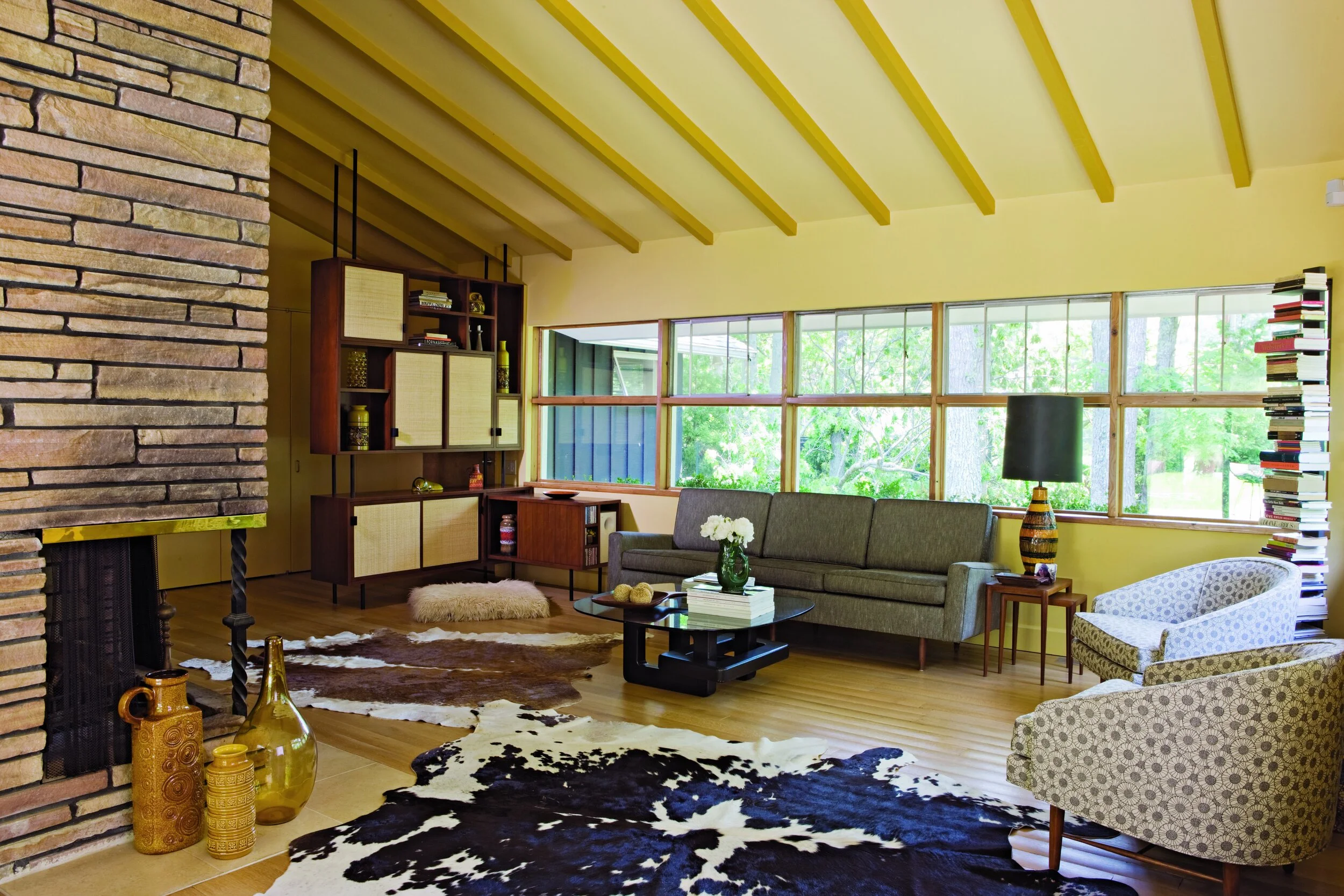 Living room with yellow walls and ceiling beams, large window with view of trees, mid-century modern furniture, stacked books, cowhide rug, fireplace, decorative vases, and a floor lamp.