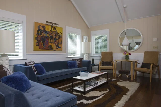 Living room with beige walls, three windows with white blinds, a dark blue sofa with pillows, a wooden coffee table with books, two beige armchairs, a round mirror on the wall, a white radiator, a flower vase on a small table, and colorful artwork on