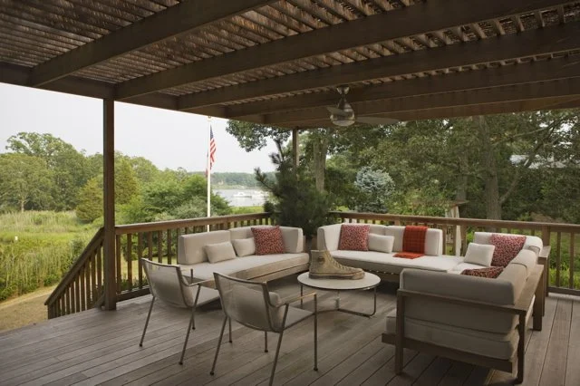 Outdoor covered patio with white sectional sofa, chairs, and a coffee table, overlooking a lush green landscape with trees and an American flag in the background.