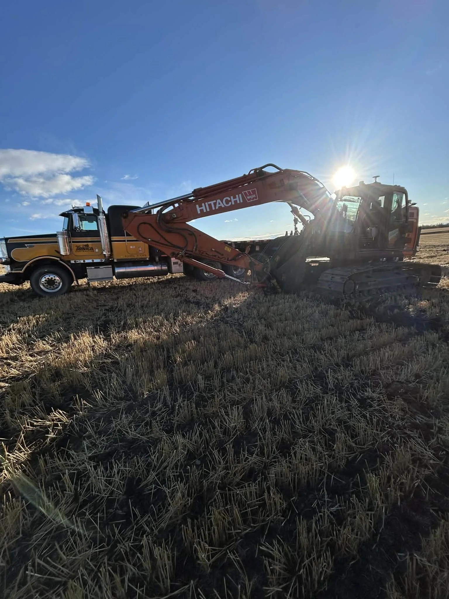 A red and black Hitachi excavator working on a field with dry grass, with the sun setting or rising in the background and a blue sky with a few clouds.