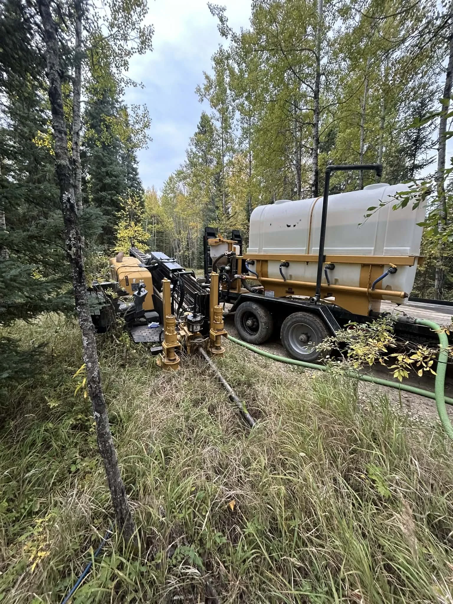 A large drilling machine set up in a forested area with tall trees and grass, connected to a water tank, preparing to drill underground.