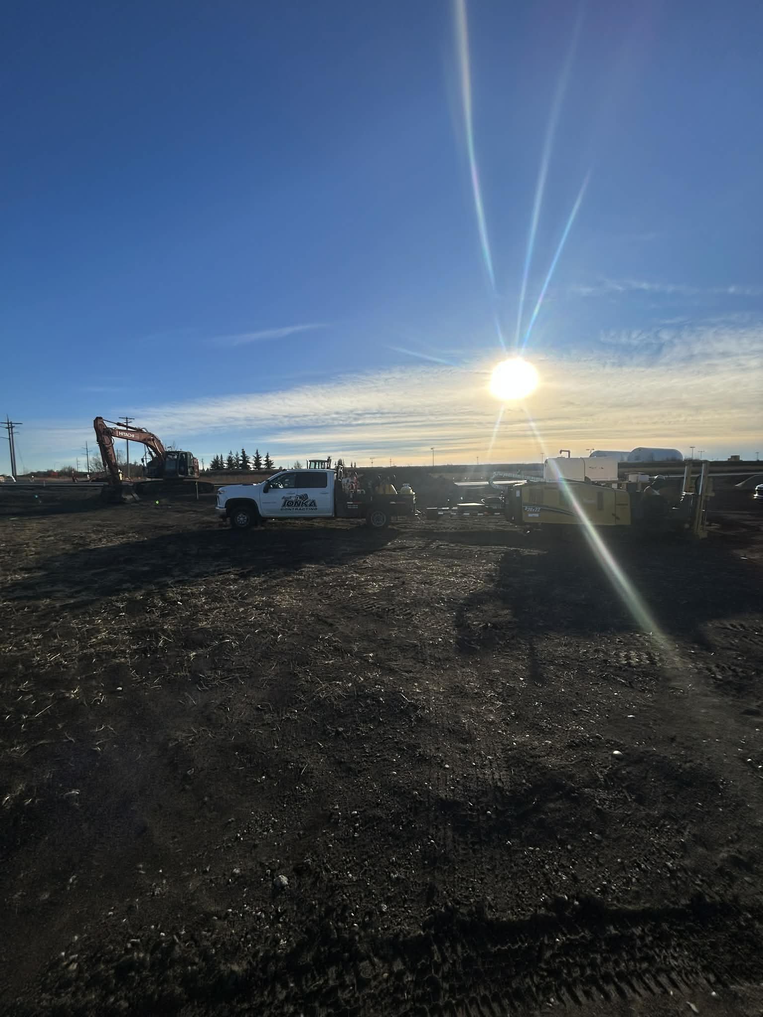 Construction site during sunset with construction equipment, trucks, and machinery on dark dirt ground.