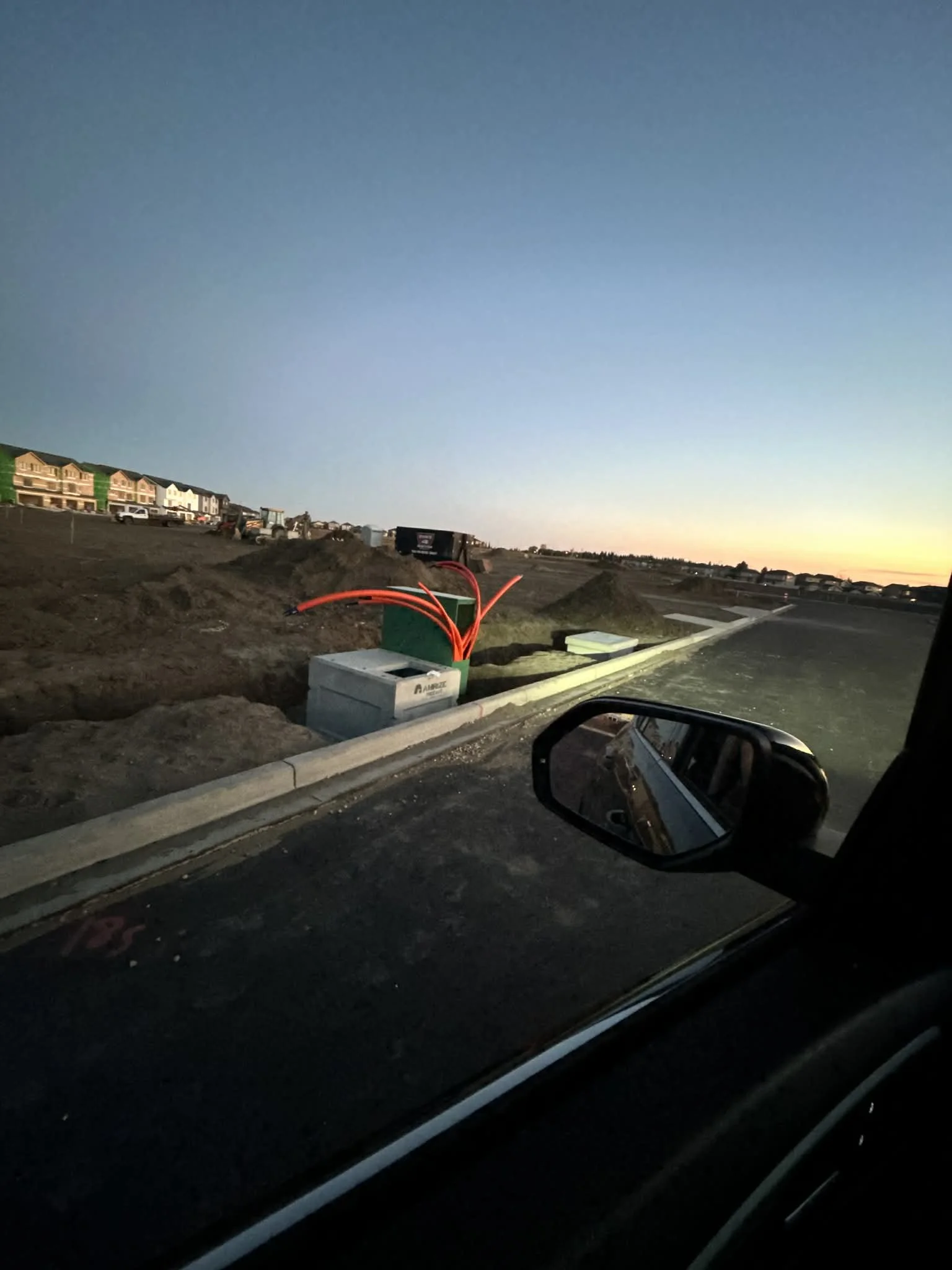 Construction site with electrical equipment and wiring along a curb during sunset, with houses in the background.