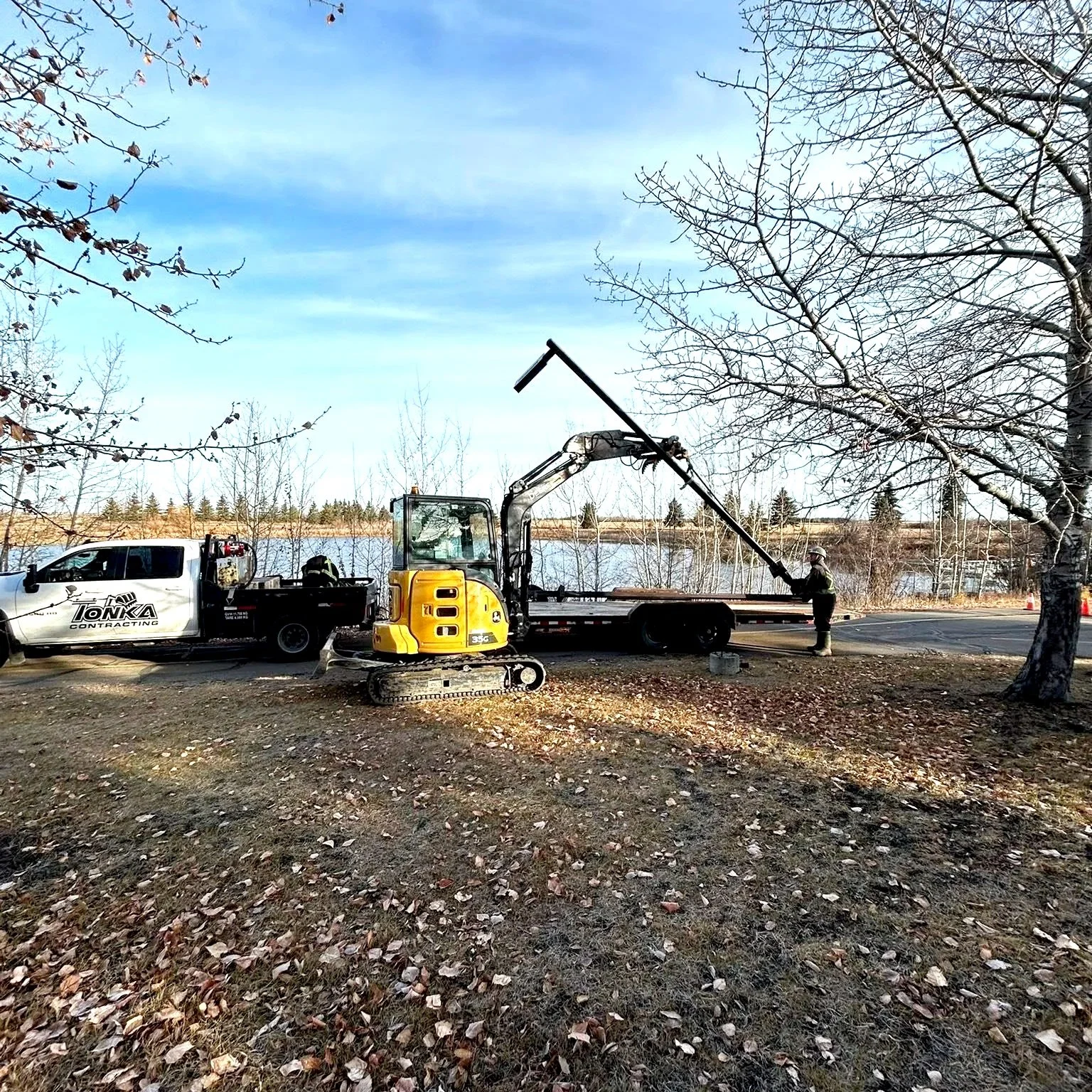 Construction workers using a compact excavator to lift a long pipe near a truck on a patch of ground with fallen leaves, trees, and a body of water in the background.