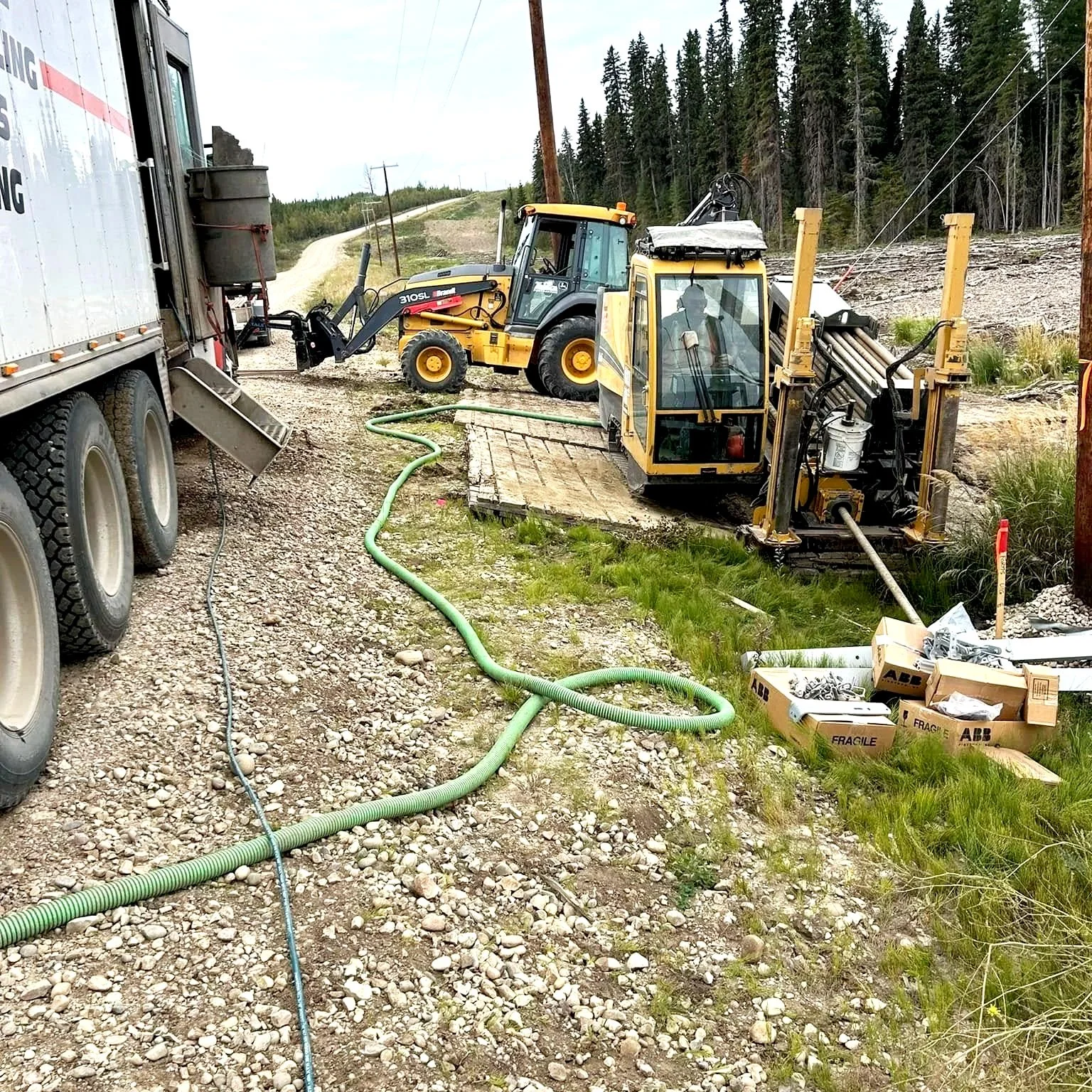 Construction vehicles working on a road, with a large truck on the left, a small excavator in the middle, and a compact track loader on the right, surrounded by gravel, grass, and trees in the background.