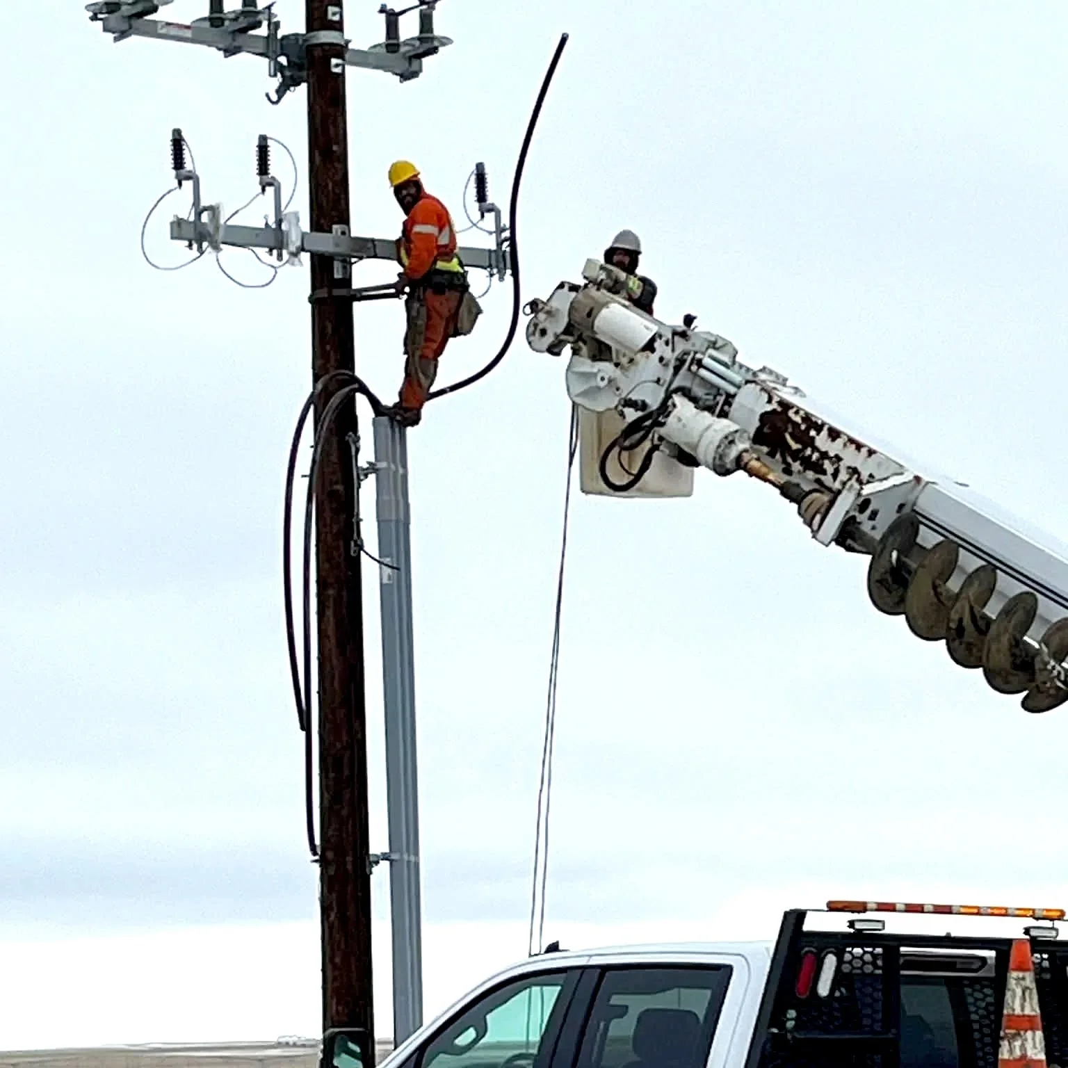 Two utility workers, one in orange safety gear and the other in white, are working on power lines using a bucket truck and climbing the utility pole.
