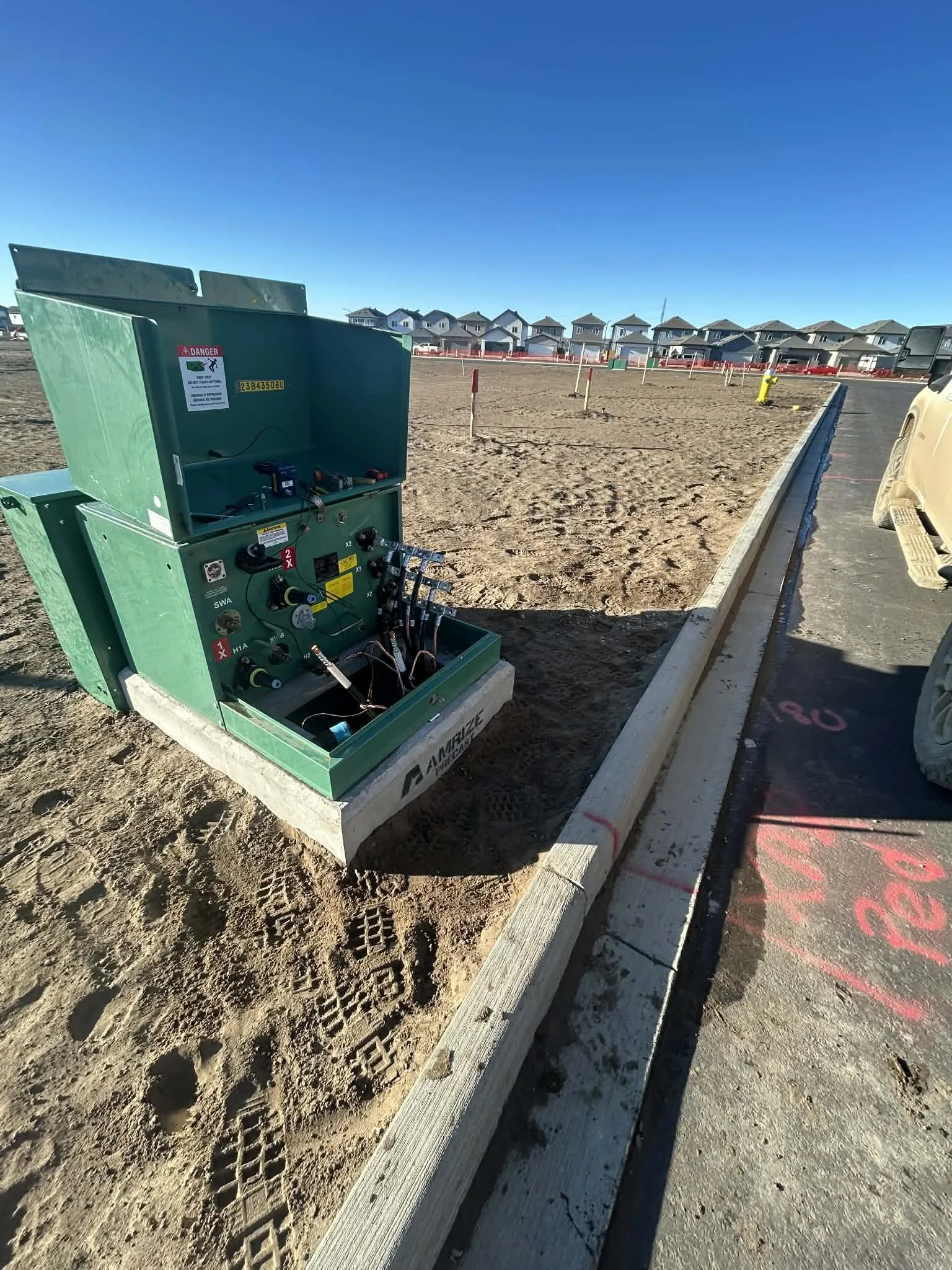 Electrical underground utility box on construction site next to sidewalk with sand, tire tracks, and footprints, with a row of houses in the background under a clear blue sky.