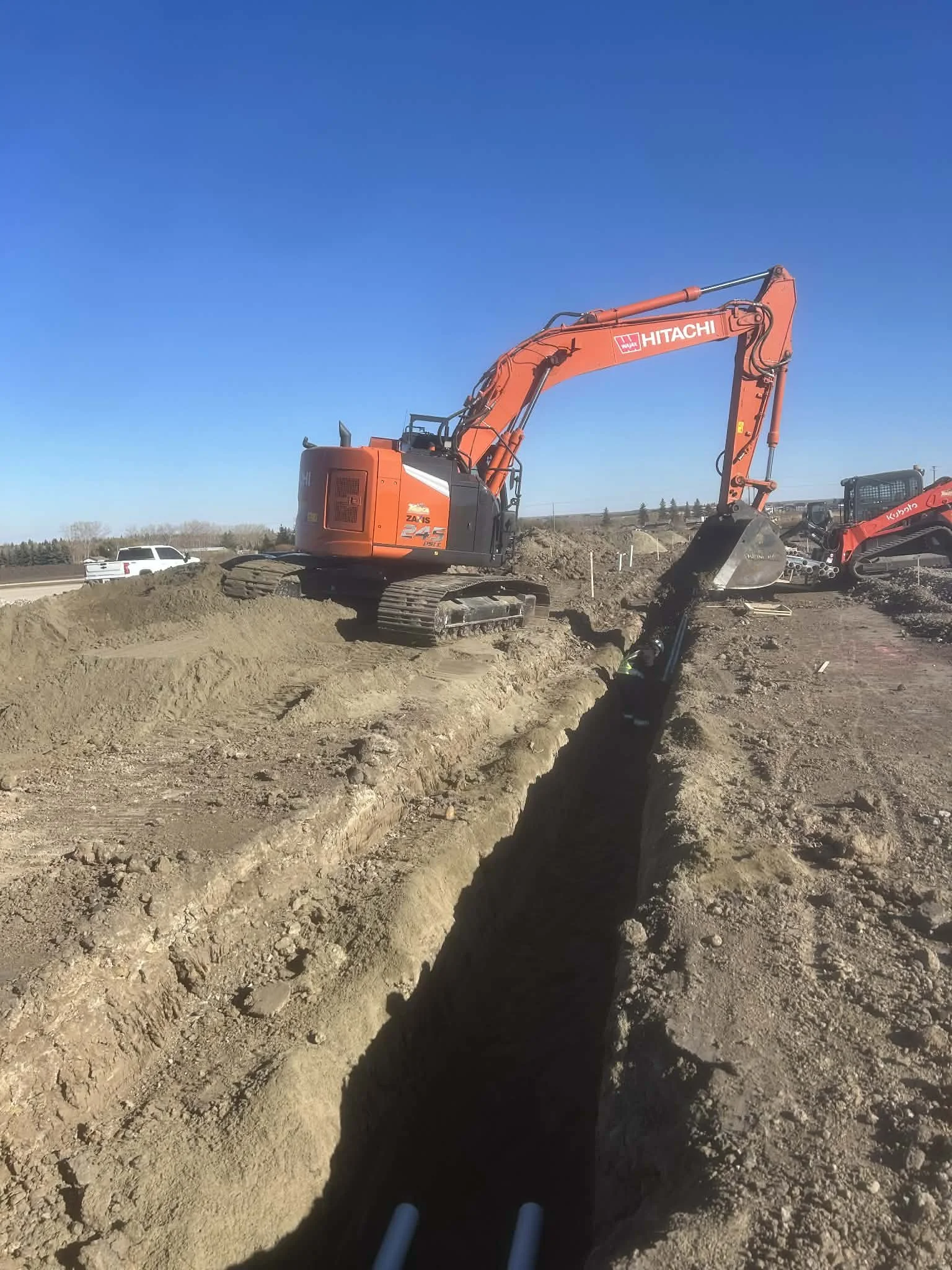 Construction site with an orange Hitachi excavator digging a trench, blue sky, other construction equipment nearby, and vehicles passing in the background.