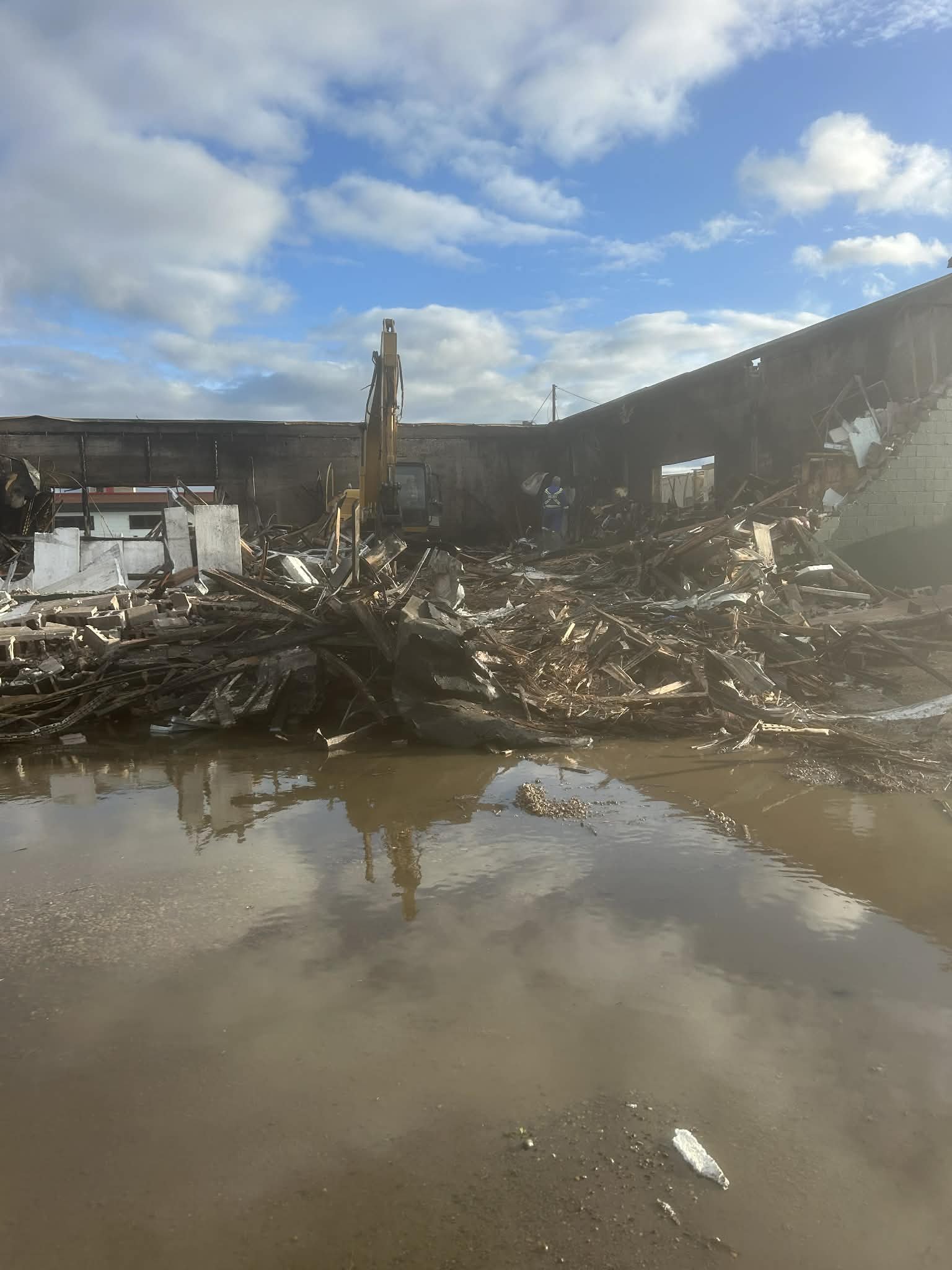 Remains of a building after destruction with debris and rubble, a construction vehicle, and reflective water on the ground under a partly cloudy sky.