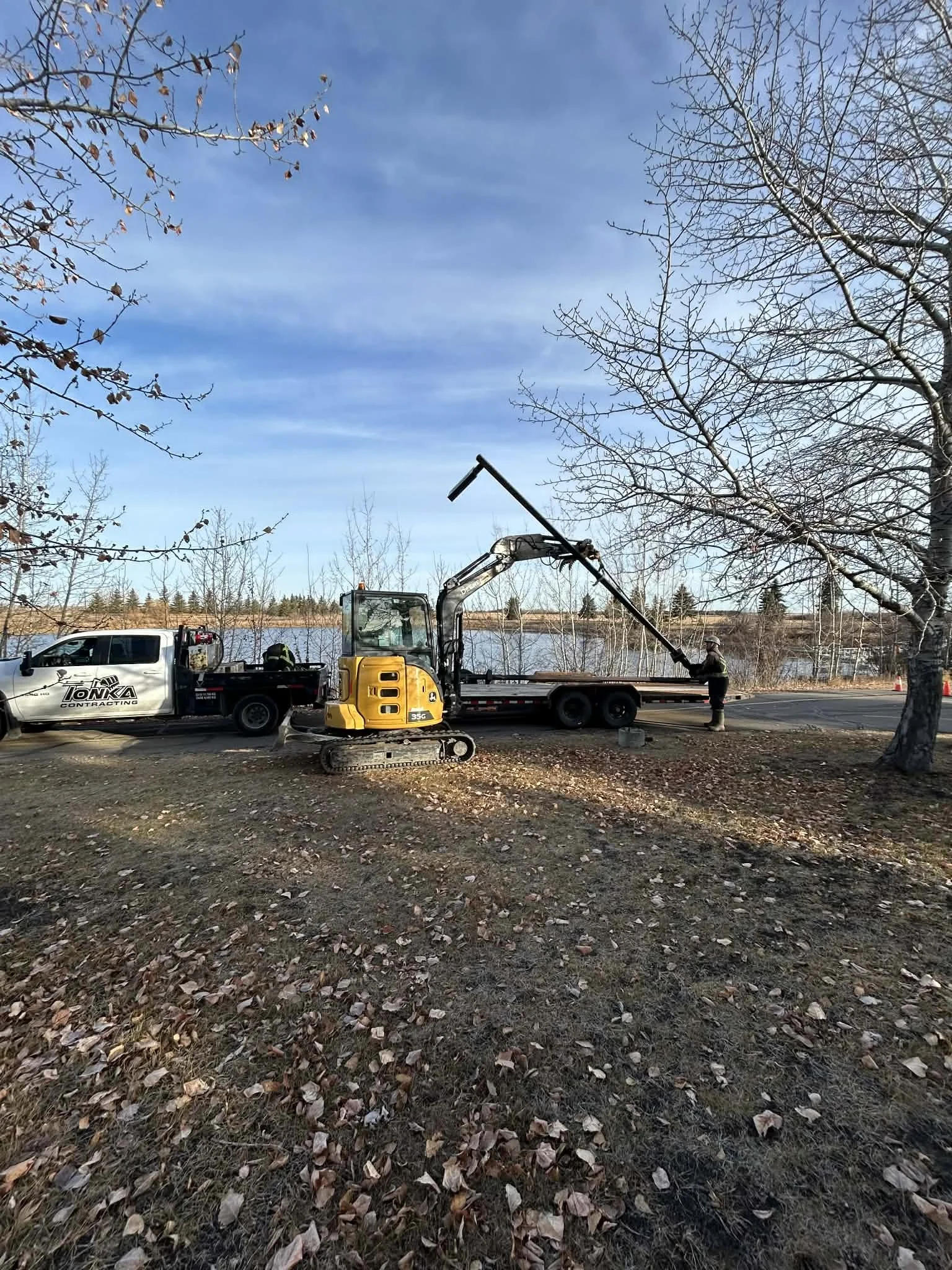 A construction scene with a small yellow excavator on tracks, a dump truck, and two workers installing a streetlight near a body of water. Bare trees and a blue sky are in the background.