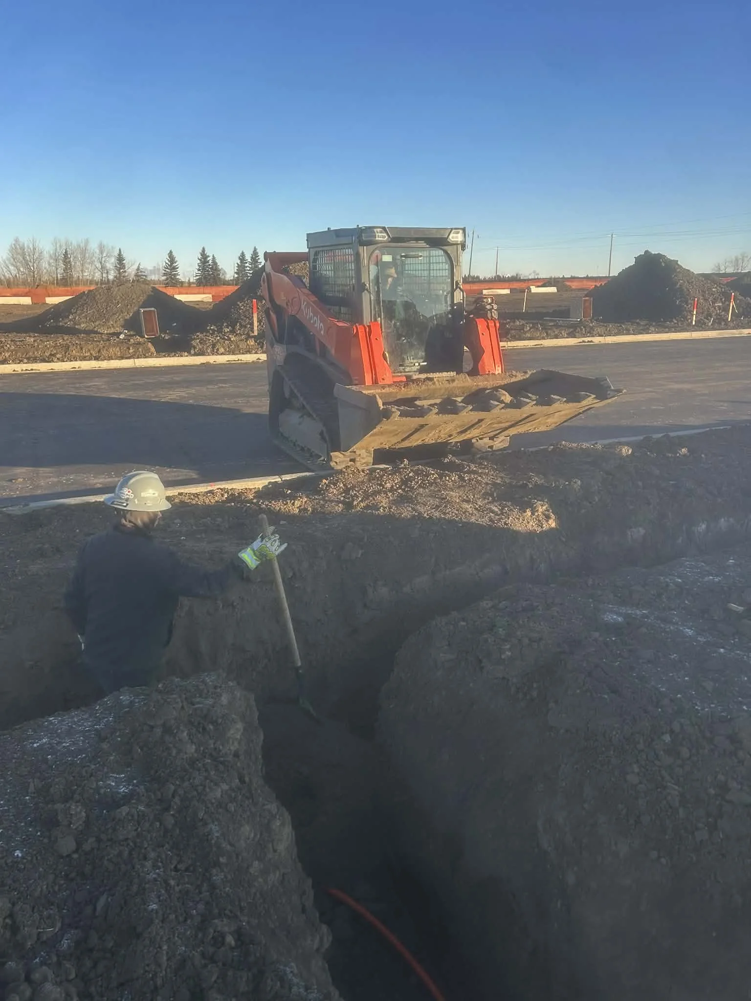 Construction worker with a hard hat and gloves standing in a trench, holding a shovel, while a small excavator operates on a nearby road construction site with piles of dirt in the background.