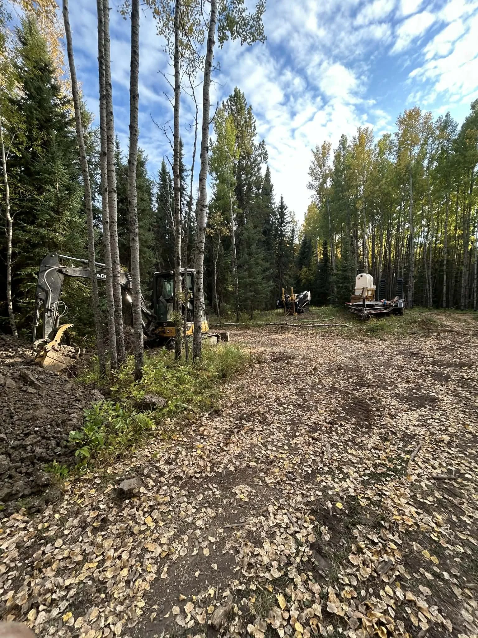 A cleared forest area with construction equipment, including an excavator, and tire tracks on the ground, surrounded by trees under a partly cloudy sky.
