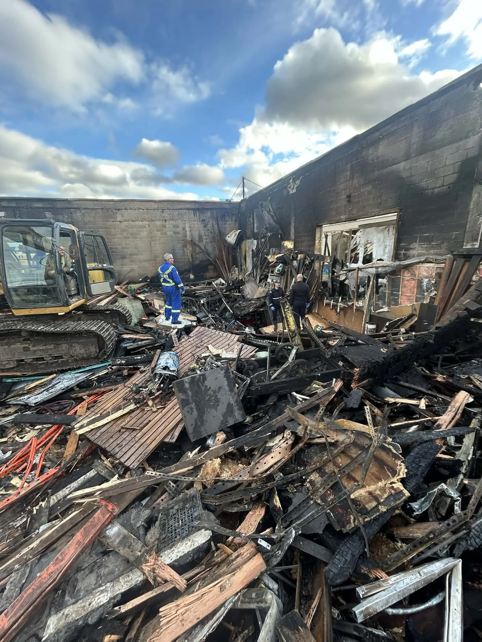 Debris and wreckage from a fire scene with blackened walls, burned materials, and a bulldozer, as people and emergency responders assess the damage under a partly cloudy sky.