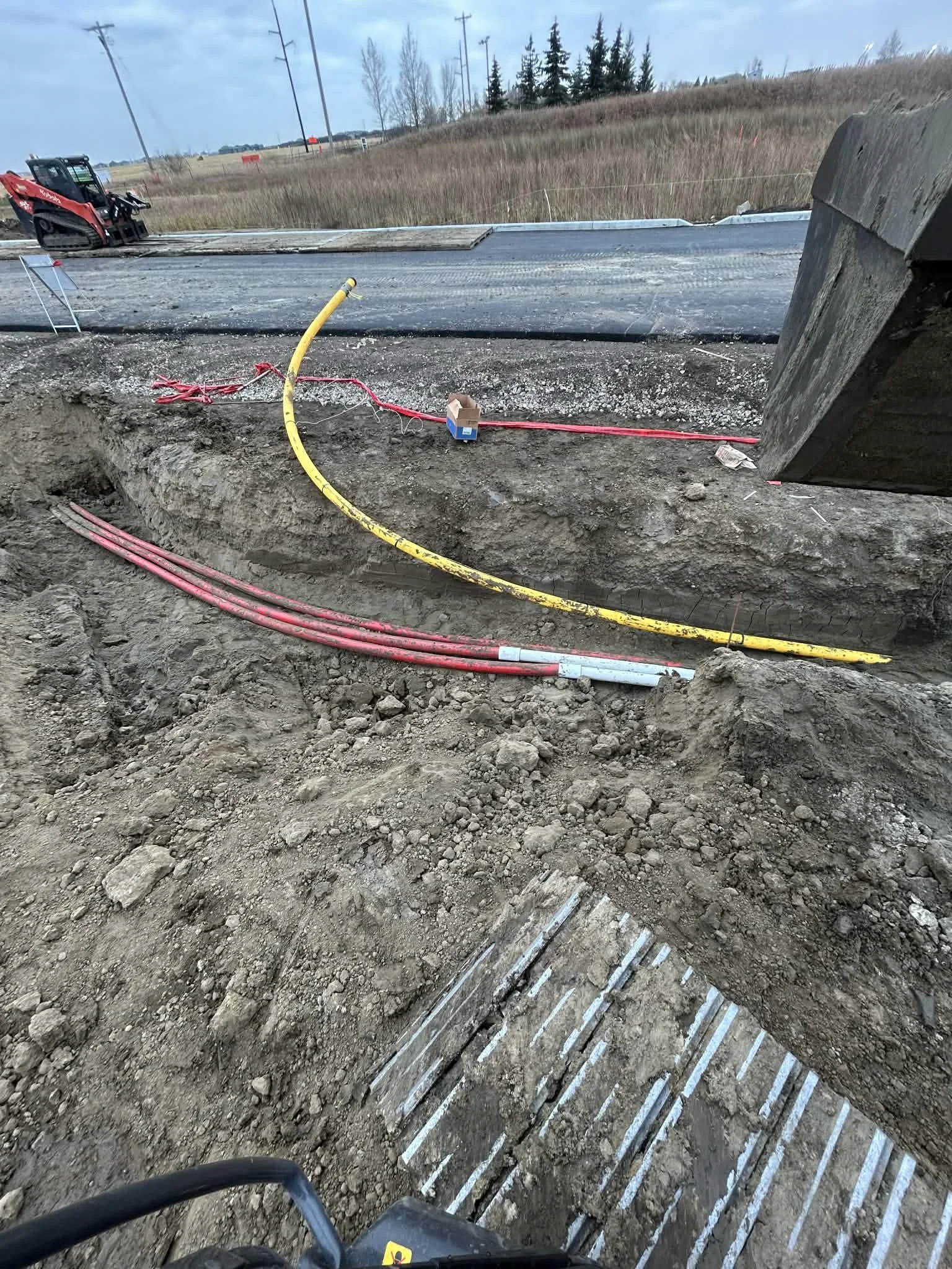 Construction site with underground electrical or plumbing pipes in a trench. Red and yellow hoses or conduits are laid in the trench, with an excavator or machine in the foreground.