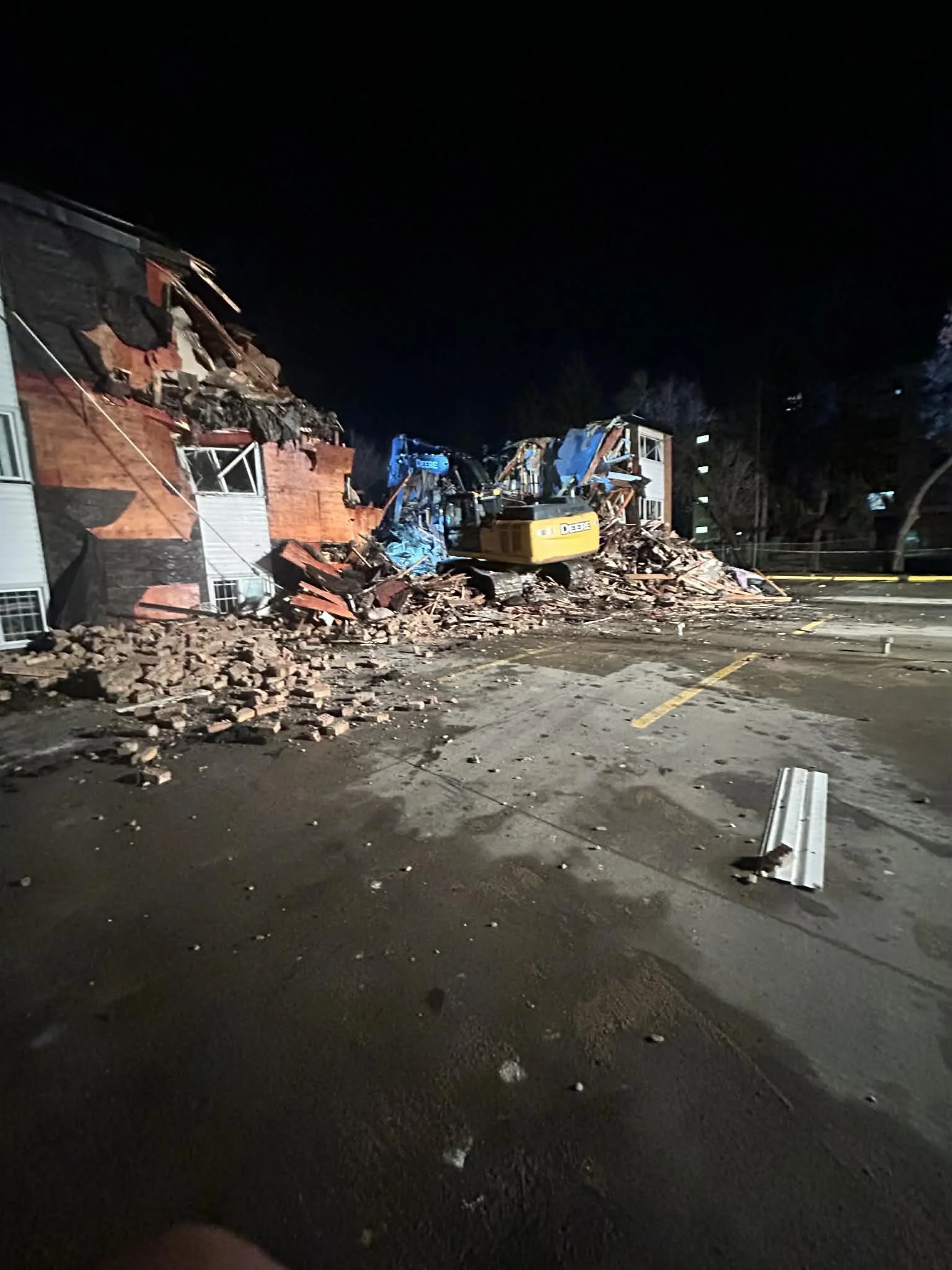 Nighttime scene of a damaged brick building with debris and rubble, construction excavator working amidst destruction, parking lot with damaged pavement and fallen metal debris.