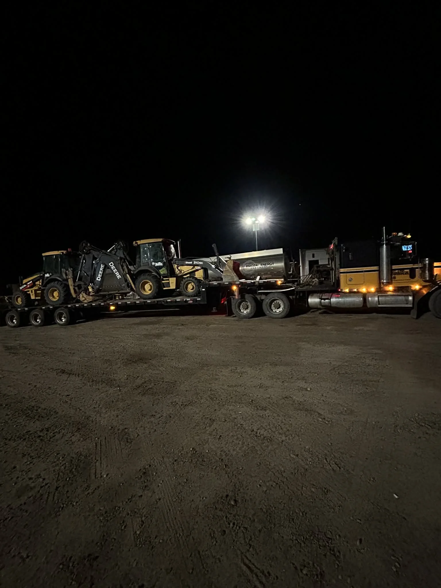 Nighttime scene with a flatbed truck carrying two John Deere construction vehicles, illuminated by bright stadium lights.