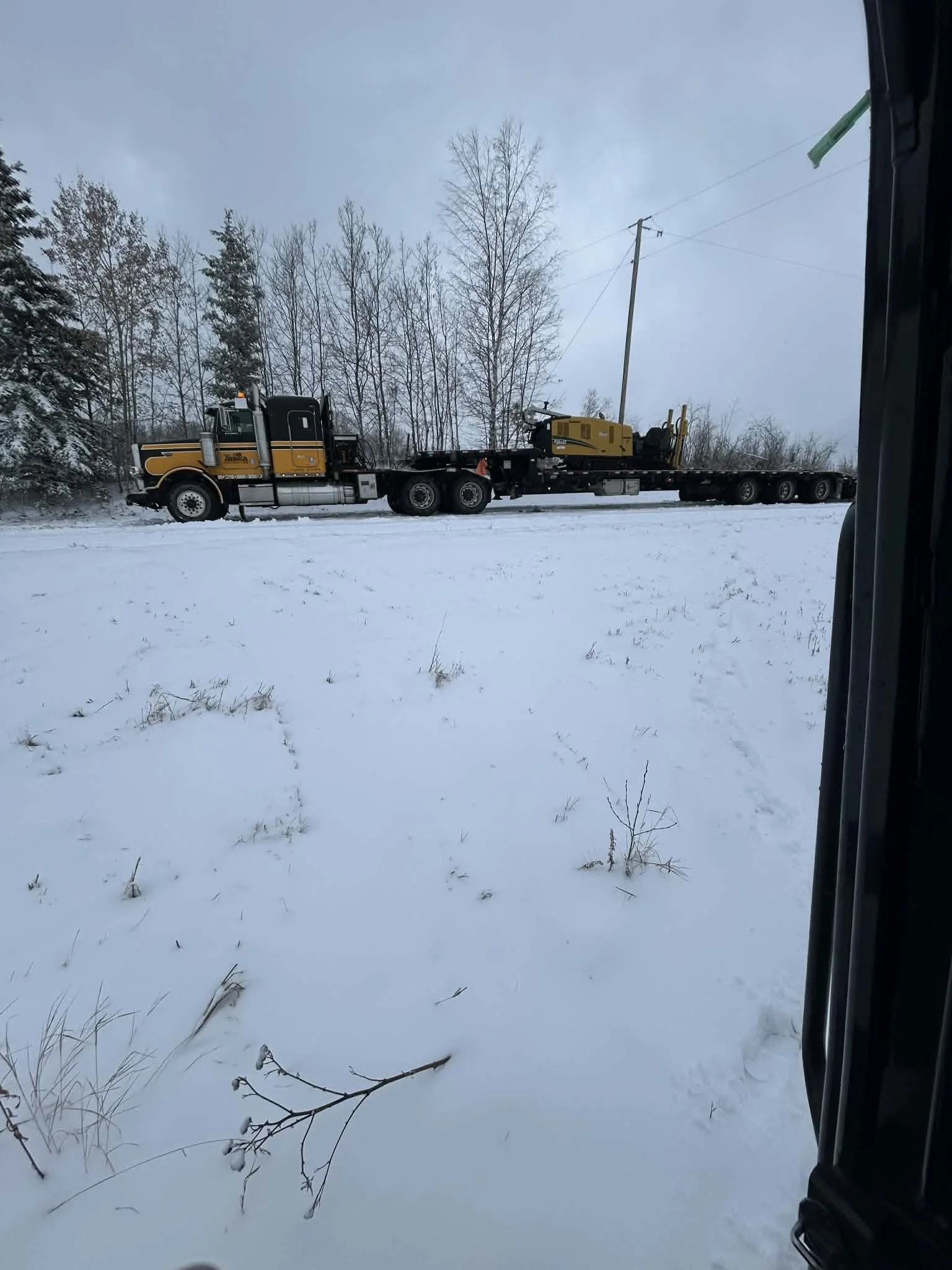 A large tow truck is pulling a yellow construction vehicle on a flatbed through a snowy landscape with bare trees and a cloudy sky.