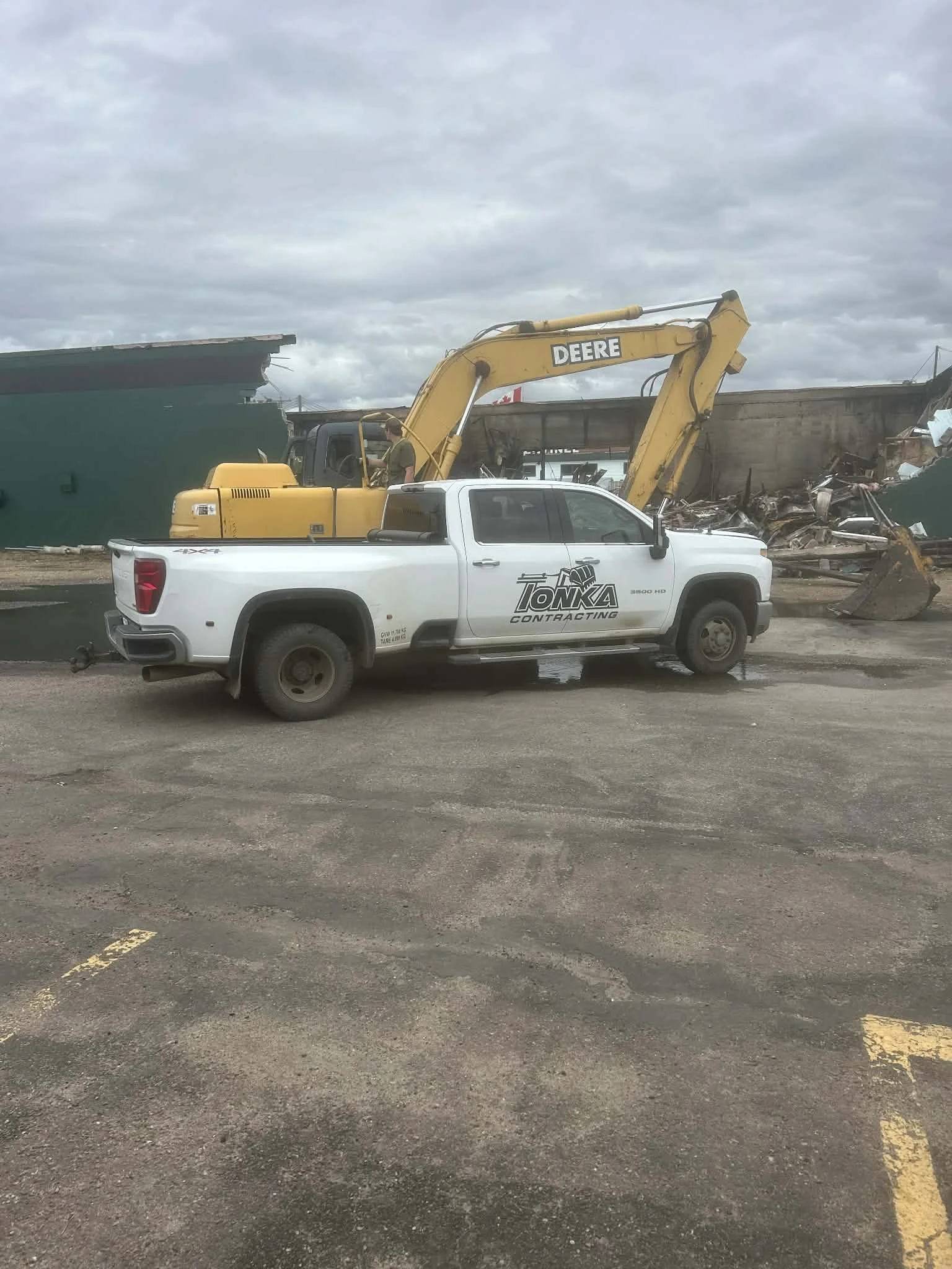 A white pickup truck with a Tonka Contracting logo and a yellow Deere excavator attached, working at a demolition site with debris and a partially collapsed structure under cloudy skies.