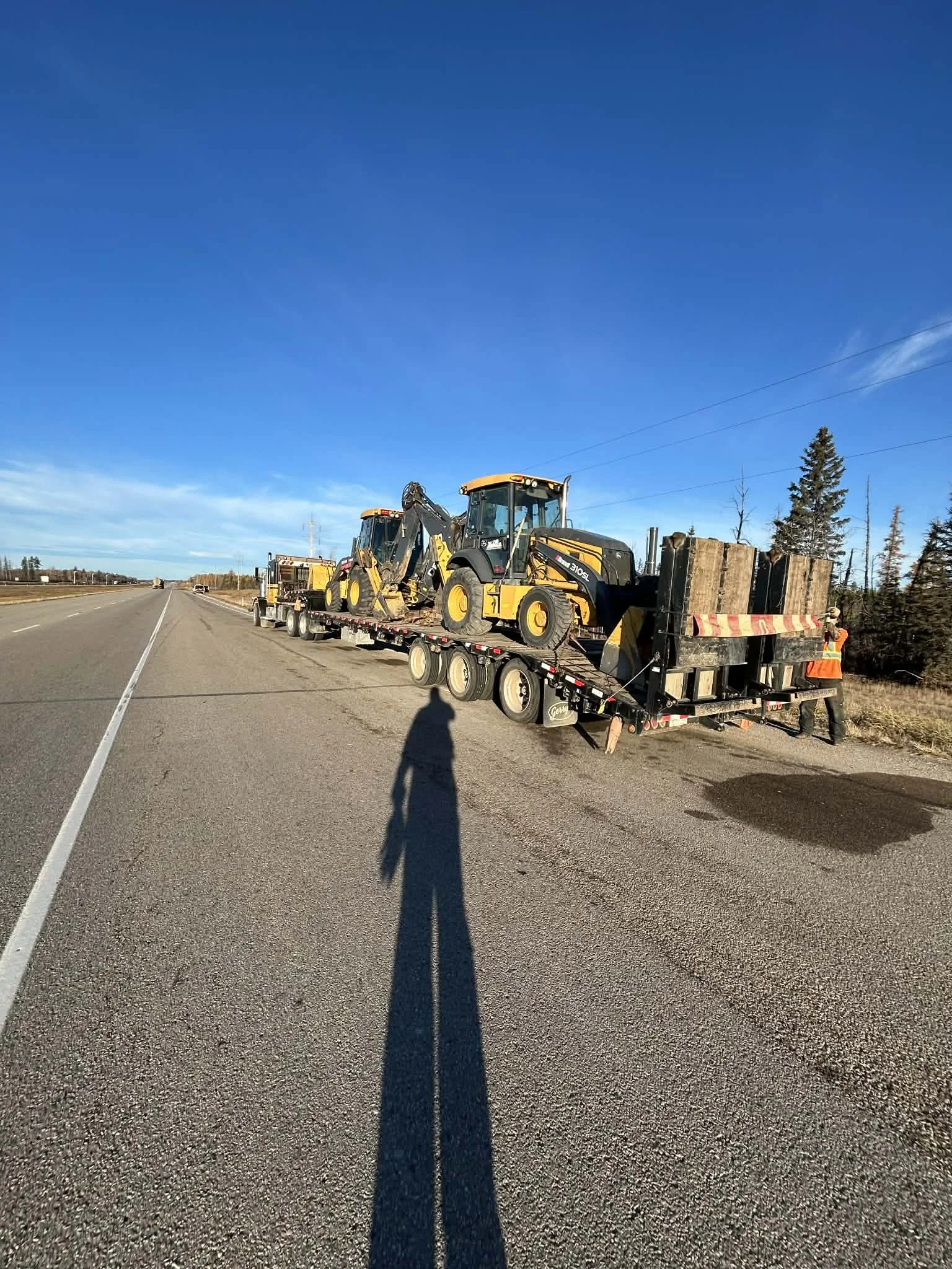 A flatbed truck carrying construction equipment, including a backhoe loader, parked on the shoulder of a highway during daytime with clear blue skies.