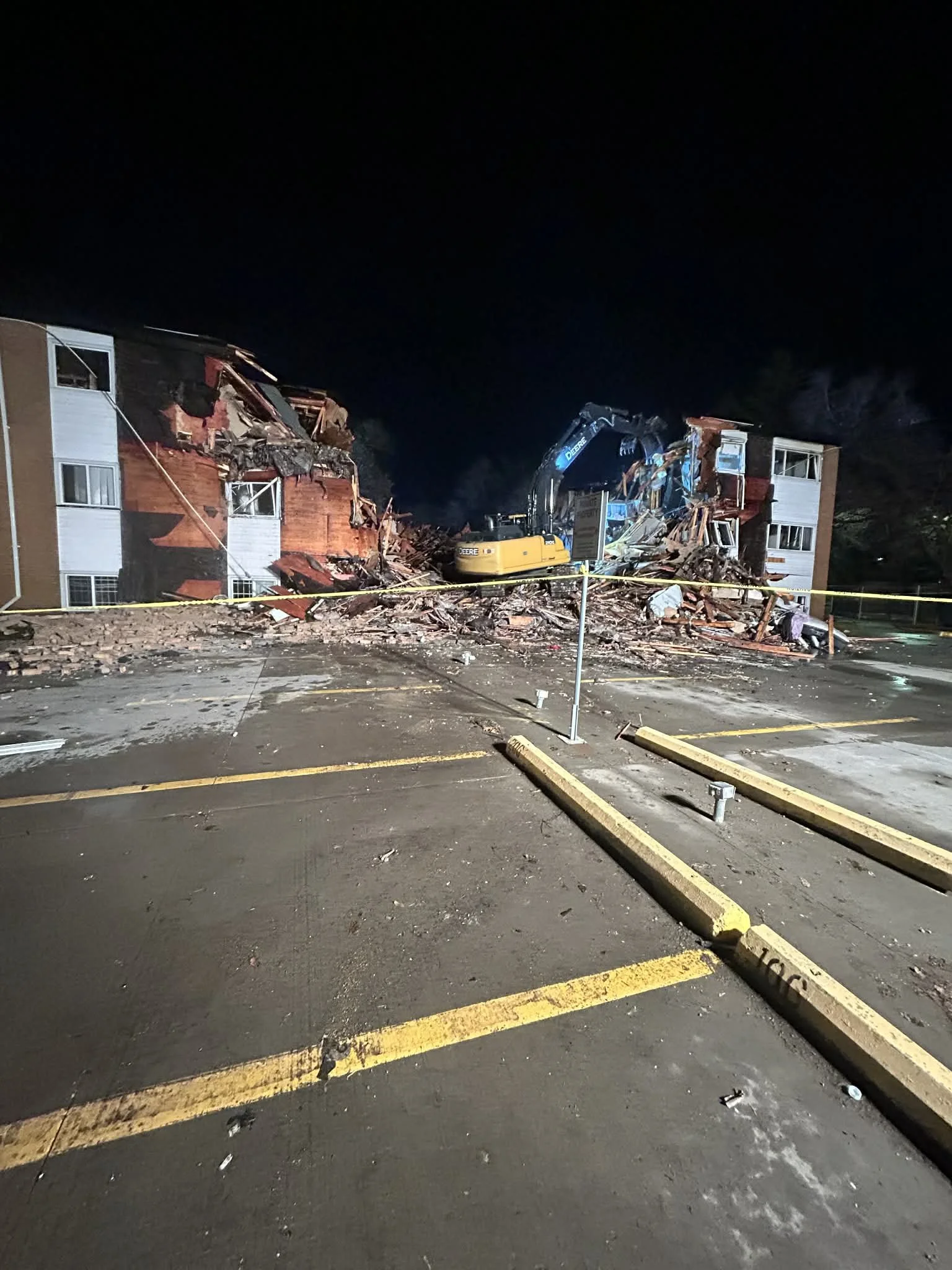 A construction excavator demolishing a section of a multi-story building at night, debris scattered on the ground, yellow parking lot bumpers in the foreground, and caution tape blocking the area.