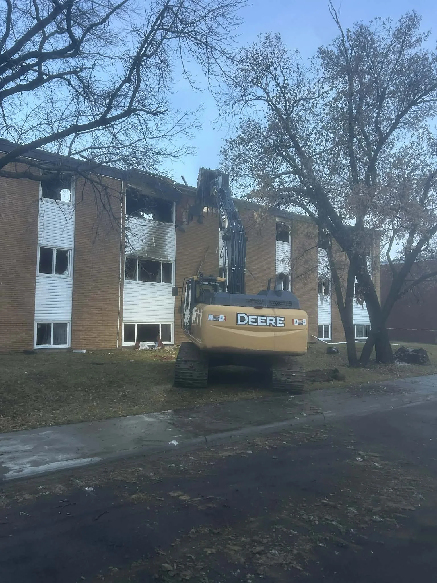 Construction excavator tearing down part of a brick apartment building, with trees in the foreground and a sidewalk in the street.