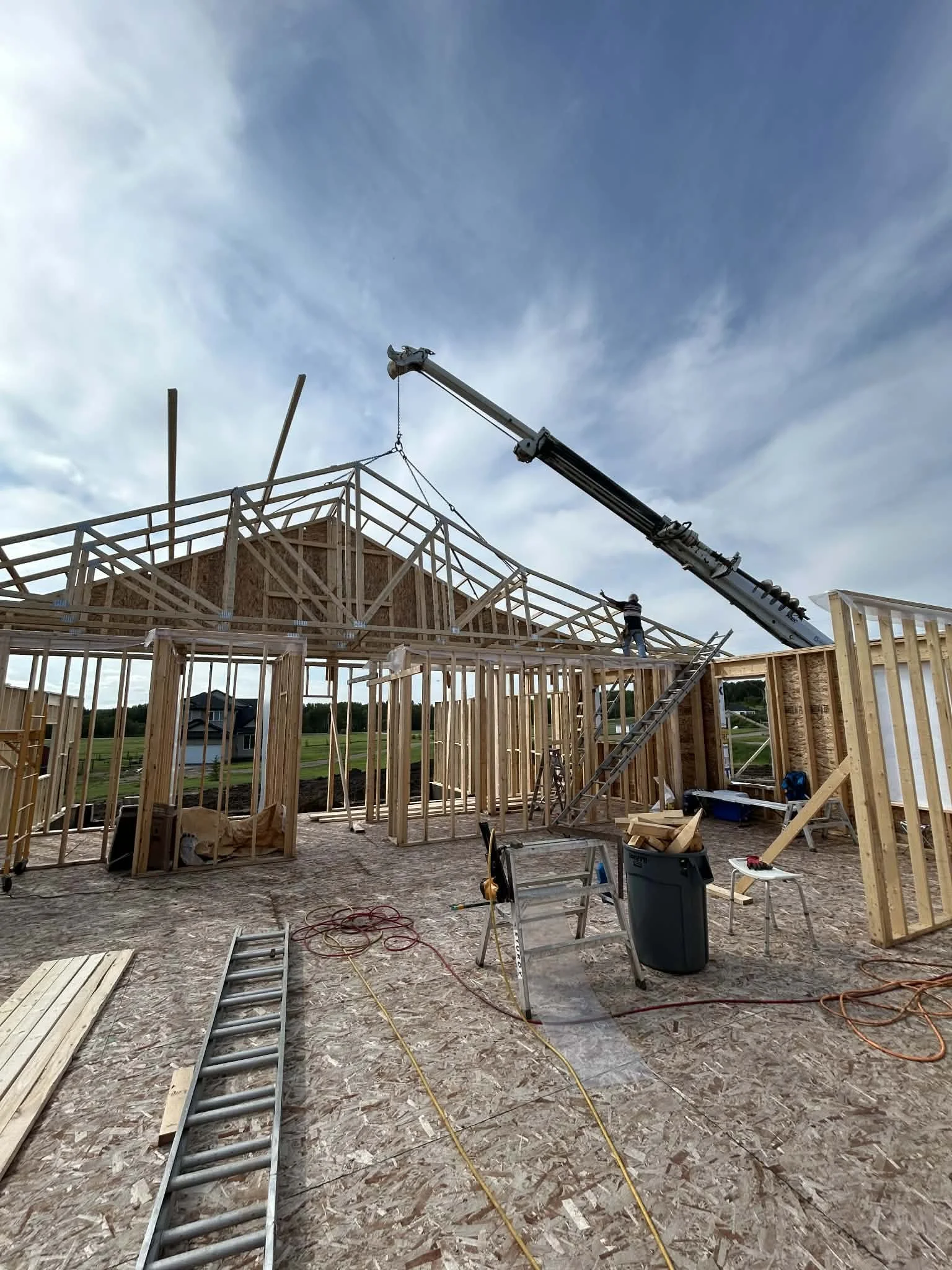 Construction workers building a wooden house frame with a crane lifting a roof truss during daylight.