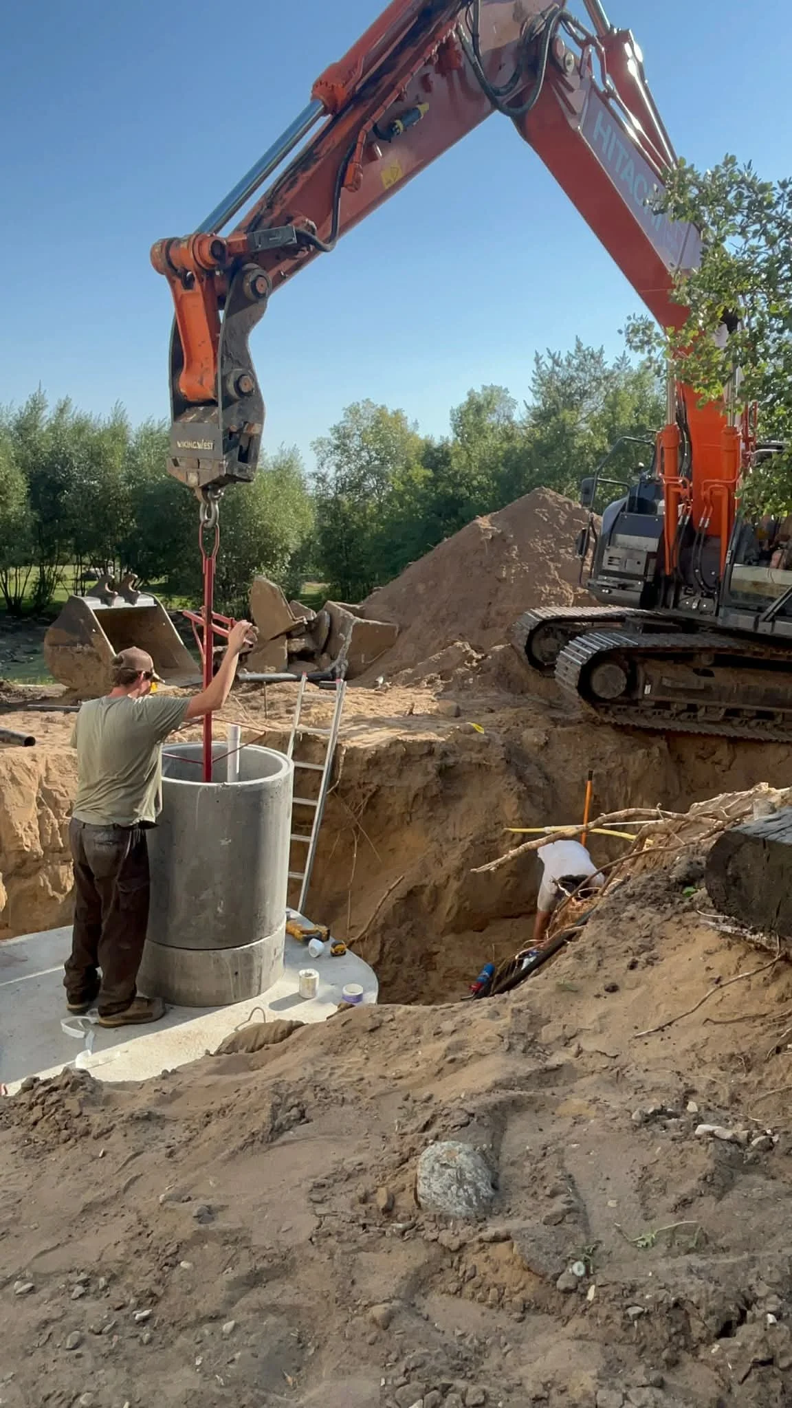 Construction site with a large orange excavator, two workers, one filling a concrete cylindrical form, and another working in a trench, with a backdrop of trees and clear blue sky.